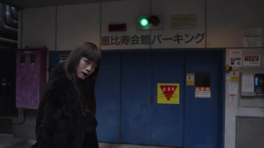 A fashionable Japanese woman with long black hair turns and looks at the camera in slow motion in a gritty urban alley