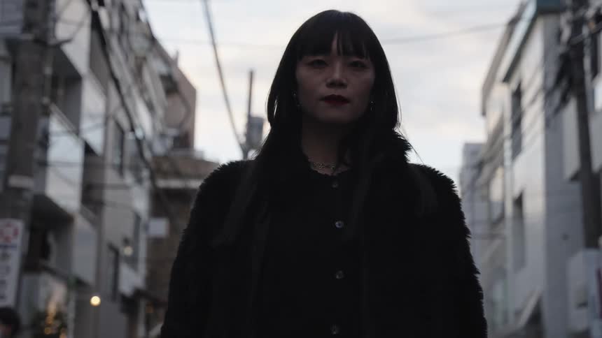 A fashionable Japanese woman in a black furry coat confidently walks toward the camera in slow motion on a Tokyo city street