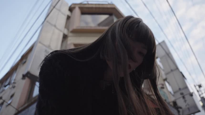 A cool young woman in a black fur coat stares down at the camera with an intense gaze on an urban street in slow motion