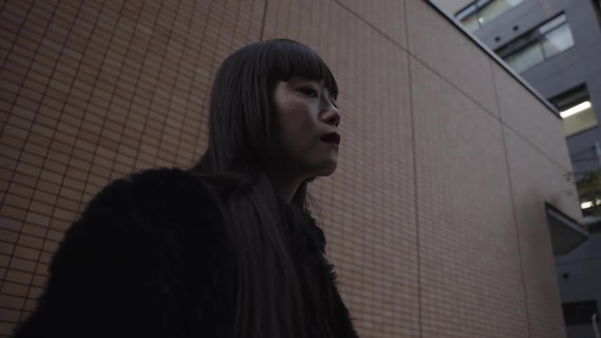 A mysterious woman with long black hair and a fur coat slowly turns to face the camera on a city street in slow motion