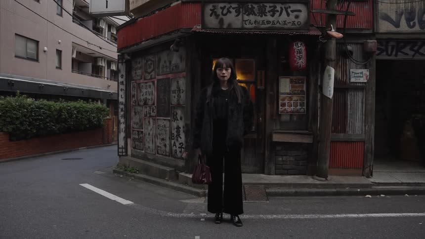 A stylish young woman dressed in black stands on a quiet street corner in front of a rustic, old-fashioned Japanese bar