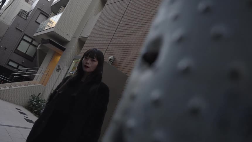 A slow reveal from behind a post shows a stylish woman with dark hair standing in a Japanese urban alley