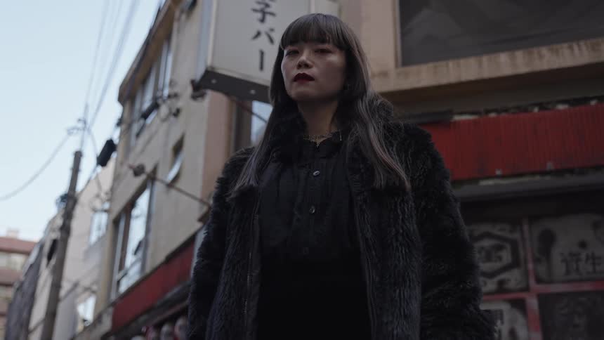A low-angle shot of a stylish Japanese woman in a black fur coat standing thoughtfully in an old urban alleyway