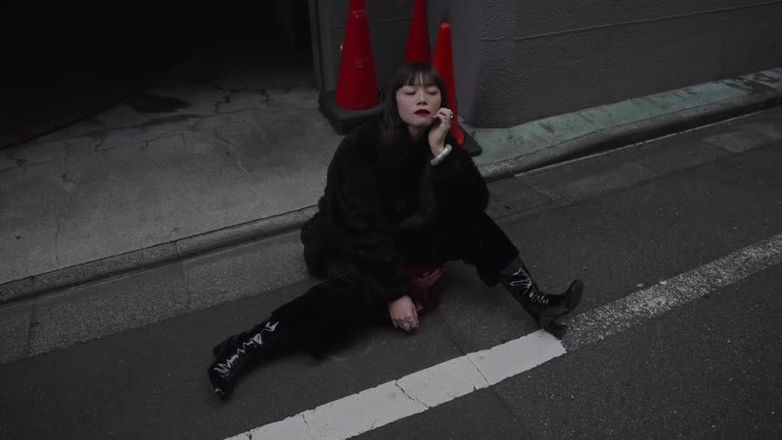 A stylish woman in a black outfit sits pensively on the pavement in a dark city alley, slowly looking up at the camera