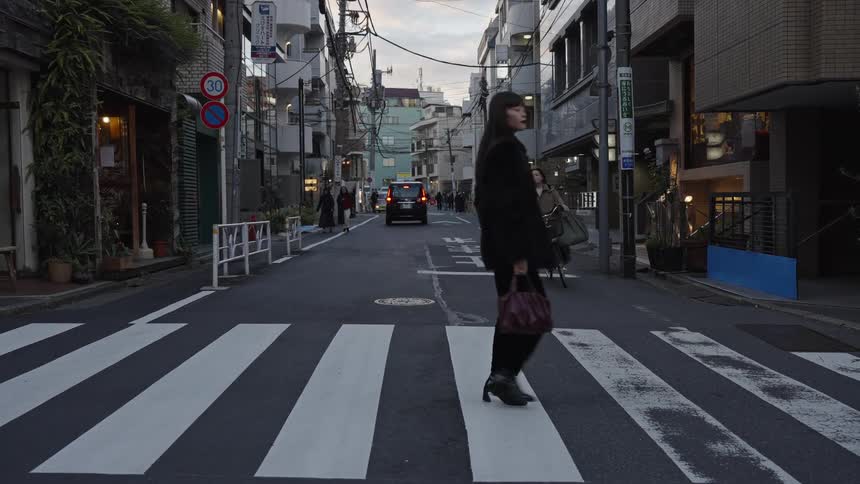 A fashionable woman walks in slow motion across a quiet city street in Tokyo at dusk