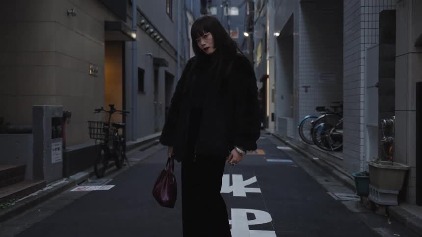 A fashionable woman dressed in all black poses with an intense gaze in a narrow urban alleyway at dusk