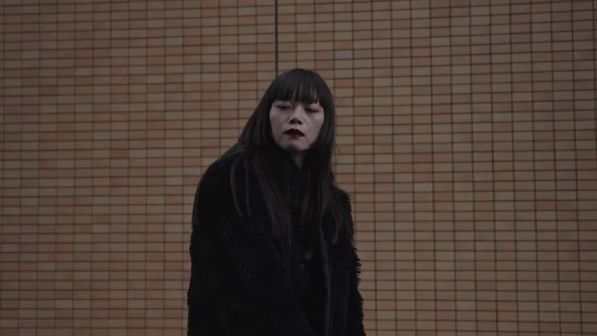 A fashionable Japanese woman with red lipstick and an all-black outfit poses against a tiled wall in slow motion