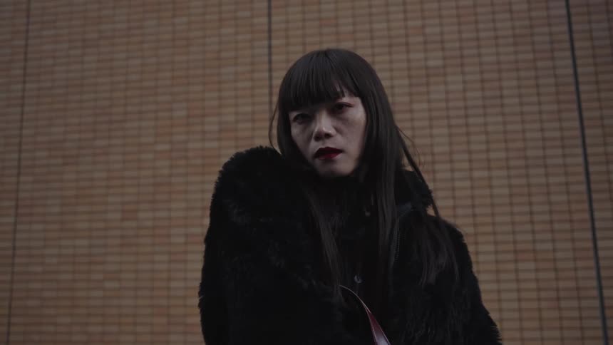 A low-angle shot of an Asian woman in a black coat looking up and staring intensely at the camera in slow motion
