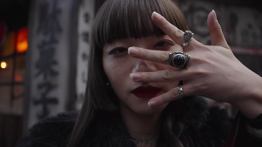 A stylish young Asian woman with bold makeup and large rings poses mysteriously on a city street, peeking through her fingers