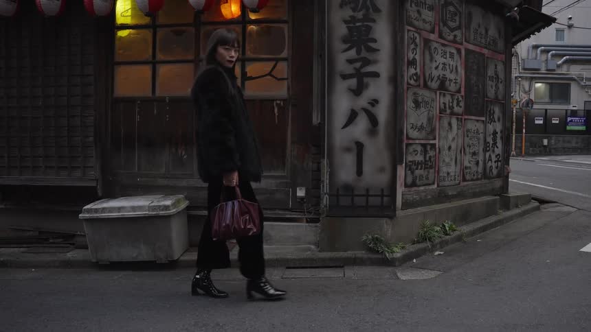A stylish woman in a black fur coat walks in slow motion across a street past a traditional Japanese bar
