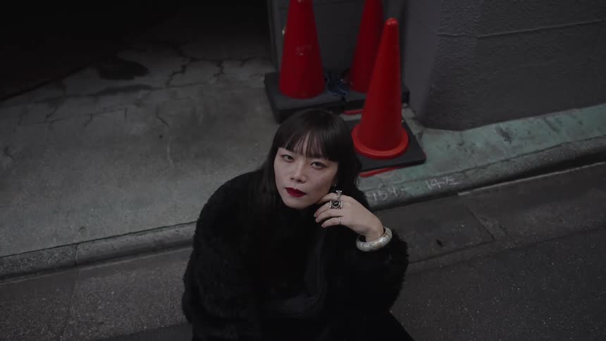 A stylish Asian woman in a black fur coat sits alone on a dark city street at night, looking up pensively at the camera