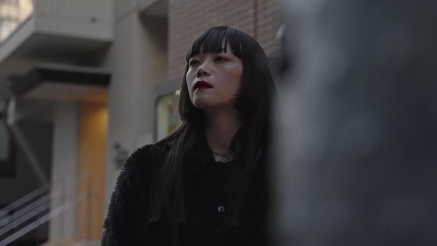 A stylish young Asian woman with red lipstick and a black coat stands pensively on a city street in slow motion