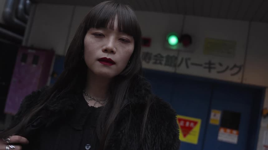 A cool Japanese woman with long black hair and red lipstick gives an intense stare directly into the camera in an urban alleyway