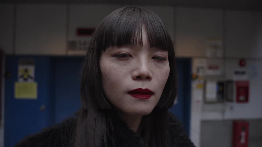 A stylish Asian woman with black hair and red lipstick looks intently at the camera as it slowly zooms in for a close-up portrait