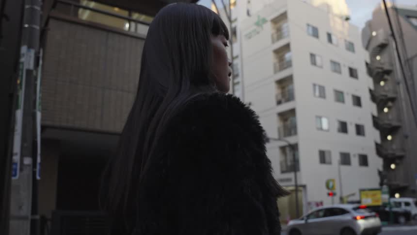 A fashionable woman in a black fur coat walks across a city street in slow motion, with the camera tilting up from her shoes