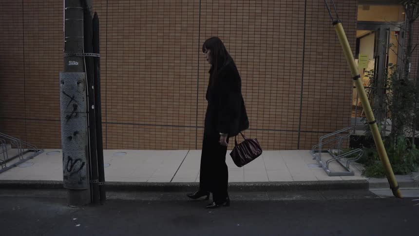 A stylish woman in an all-black outfit stands pensively on a city street before slowly turning her head away from the camera