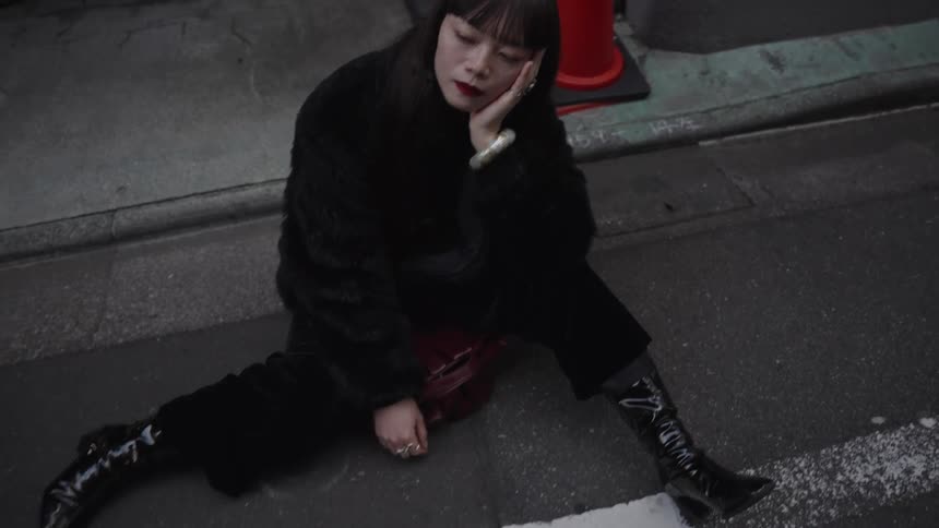 A sad and fashionable woman with red lipstick sits with her eyes closed on the pavement of a city street in a pull-back shot