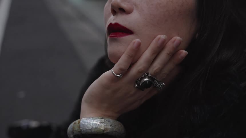 Close-up of a pensive woman with red lipstick resting her hand, adorned with bold jewelry, on her cheek