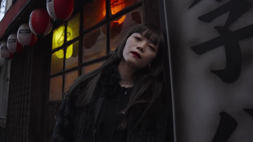 A stylish young Asian woman poses with a pensive expression against a wall in a moody Japanese alley at night