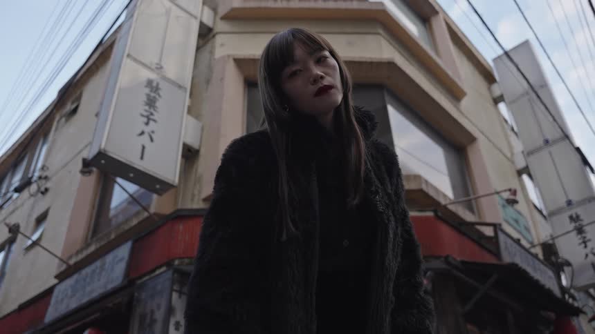 A stylish Japanese woman with an intense gaze stands on a gritty urban street corner in an all-black outfit
