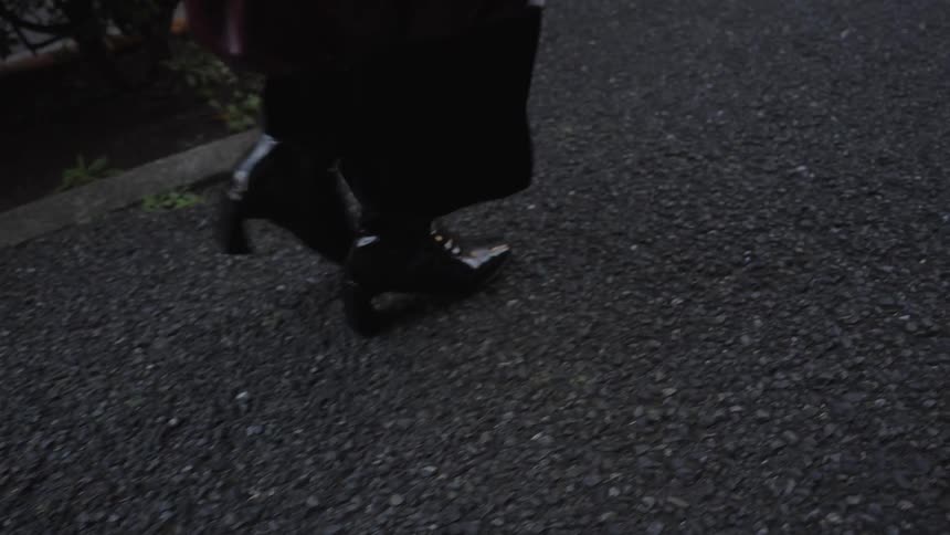 A slow-motion close-up follows the stylish black heeled boots of a person walking along a city street at dusk