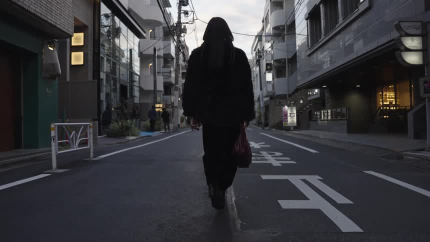 From a low angle, a woman in a black fur coat and heeled boots walks down the middle of a quiet urban street in Japan
