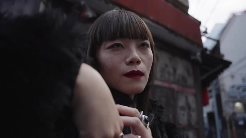 A moody, low-angle close-up of a stylish Asian woman with red lipstick and silver rings standing in a city alleyway