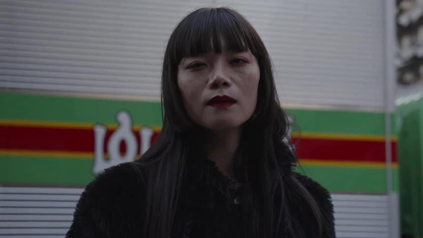 An intense close-up portrait of a Japanese woman with dark bangs and red lipstick staring directly into the camera on a city street