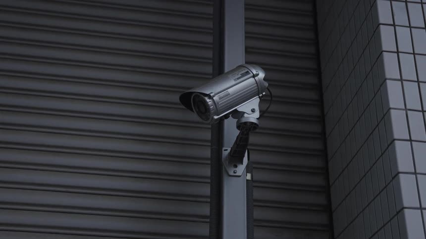 A silver security camera is mounted against a dark industrial building and roll-up door