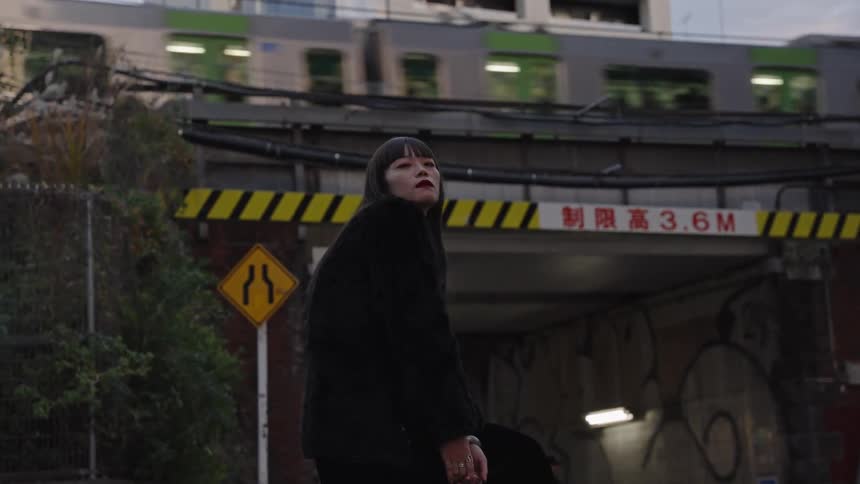 A stylish woman in all black poses on a gritty city street as a train passes overhead in slow motion