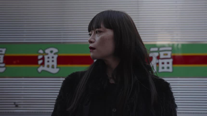 A stylish Asian woman with dark lipstick stands on a city street, slowly turning to give an intense look to the camera