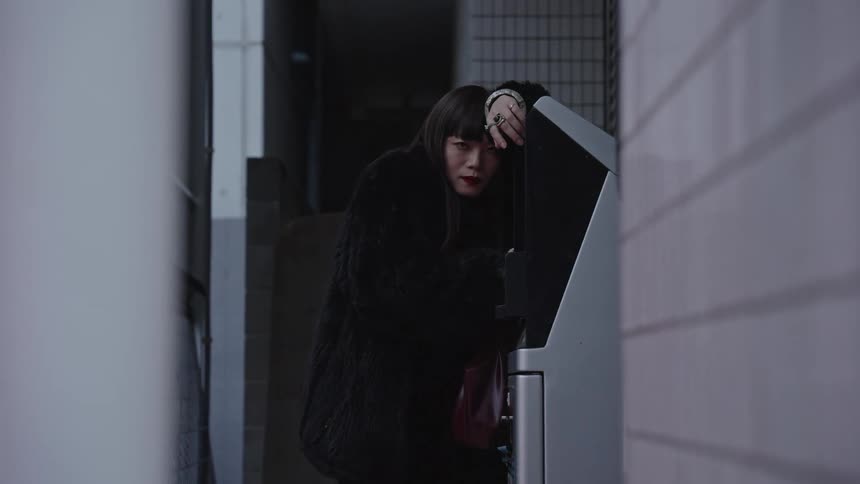 A stylish woman in a black fur coat strikes a somber pose, leaning against a machine in a dimly lit city alleyway