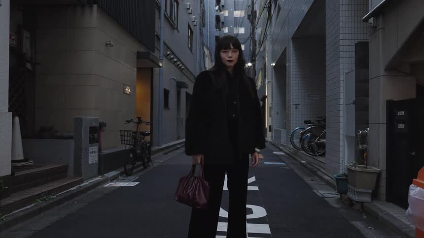 A stylish woman in all black stands and poses for the camera in a modern city alleyway at dusk