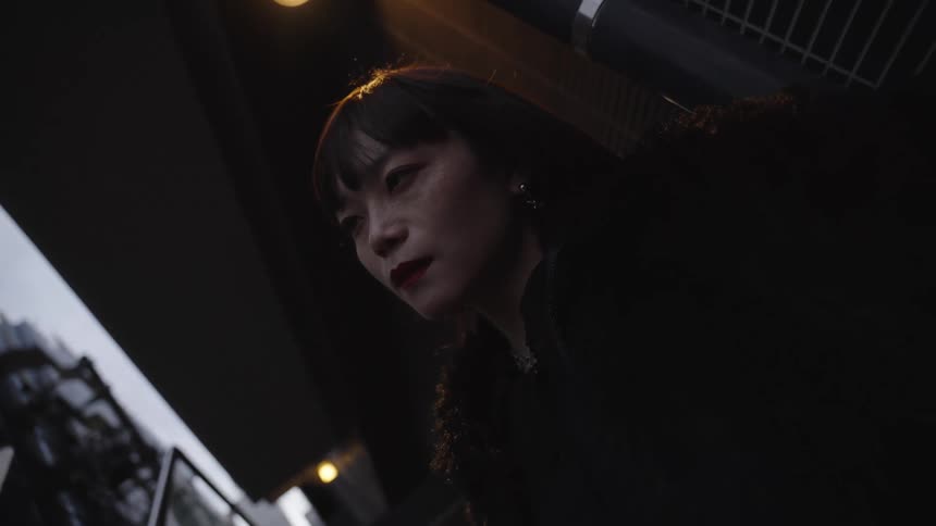 A dramatic low-angle shot captures a pensive Asian woman illuminated by a single light in a dark city environment