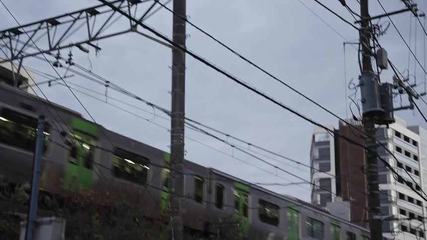A silver and green commuter train full of passengers travels through a Japanese city on an overcast day