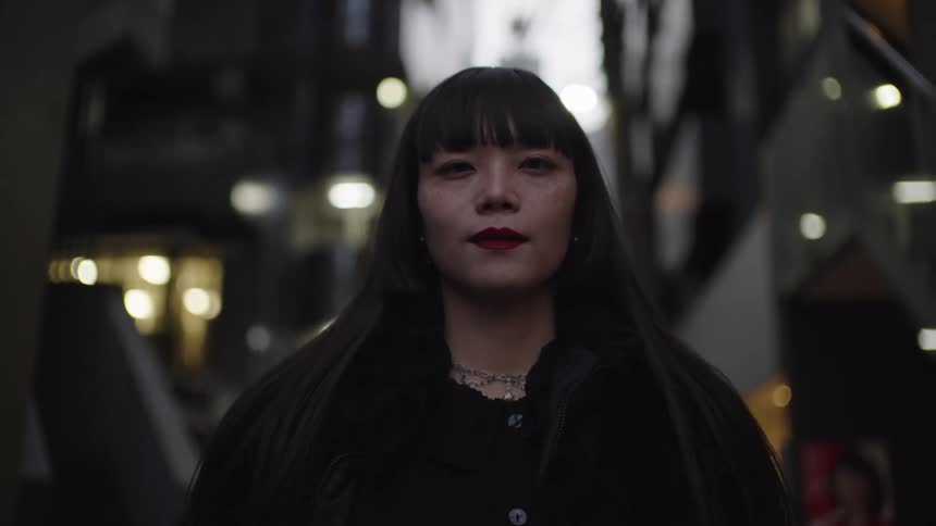 Close-up portrait of a stylish Japanese woman with long dark hair looking seriously at the camera in a modern urban environment at night