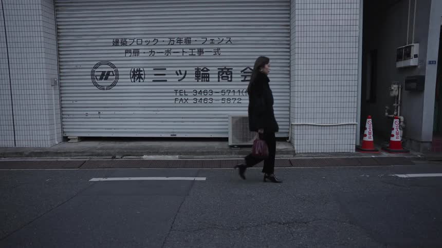 A woman dressed in black walks in slow motion across an urban street in front of a building with Japanese writing on the door
