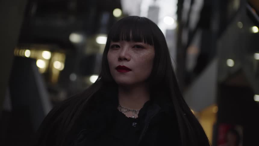 Close-up portrait of a stylish Japanese woman with dark bangs and red lipstick looking directly at the camera in a modern urban setting at night