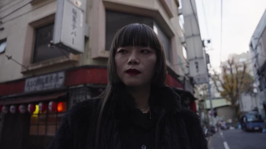 A fashionable Asian woman wearing a black fur coat stands pensively on a quiet street corner in Tokyo