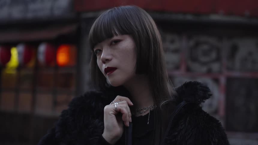 A moody close-up portrait of a stylish young Asian woman looking at the camera in slow motion on a city street at dusk