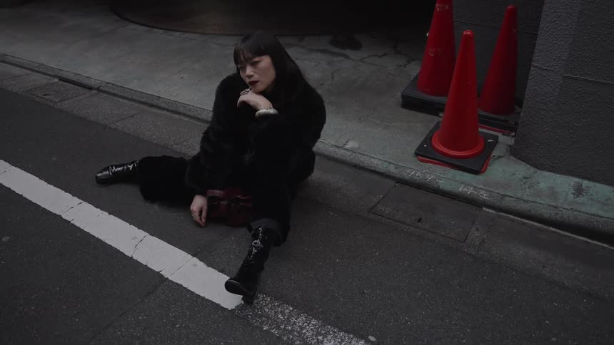 A melancholic woman in a black fur coat sits pensively on the pavement of a dark city street