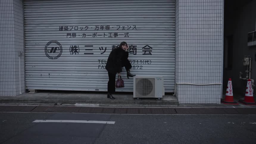 Wide shot cinemagraph of a woman in black frozen in a quirky pose against a garage door in an urban Japanese setting