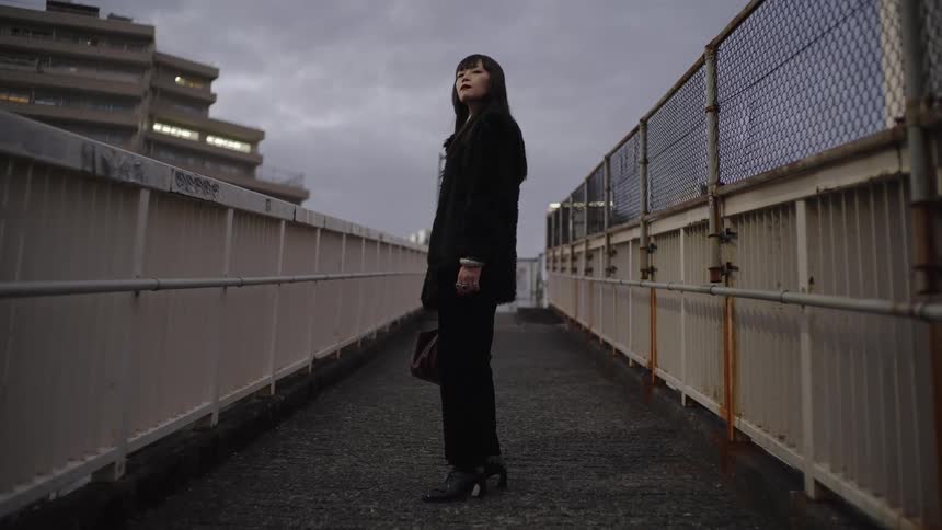 A pensive young woman in a black coat stands on an industrial walkway and looks up at the overcast sky