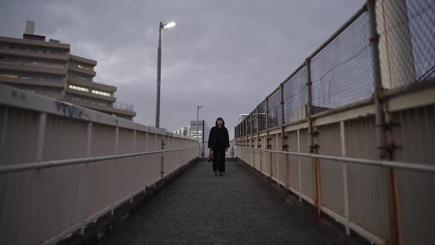A young woman stands still on a narrow, fenced urban overpass on a gray, overcast day
