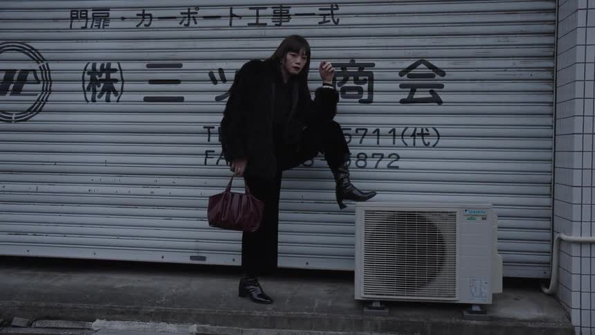 A stylish woman in a black fur coat and boots holds a cigarette and a handbag, posing on an urban street