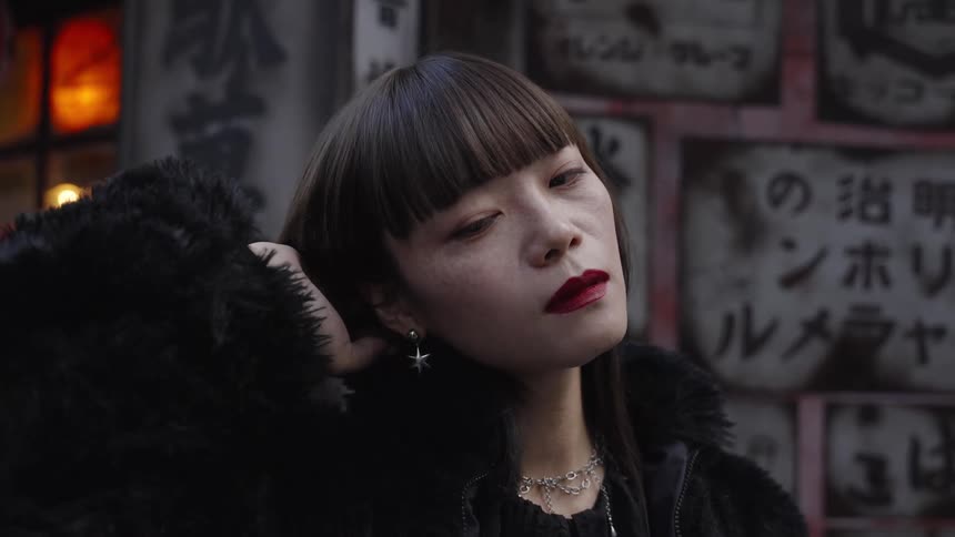 A stylish Japanese woman with red lipstick and bangs runs her hand through her hair in slow motion on a city street