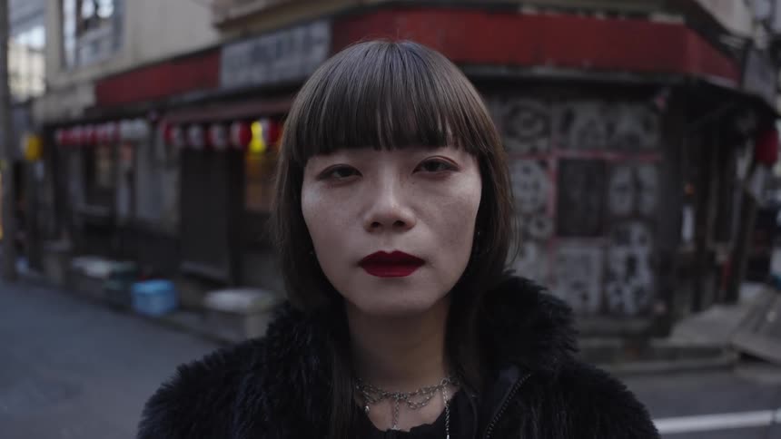 A stylish Japanese woman with an intense gaze stares directly into the camera on a city street as the shot slowly zooms out