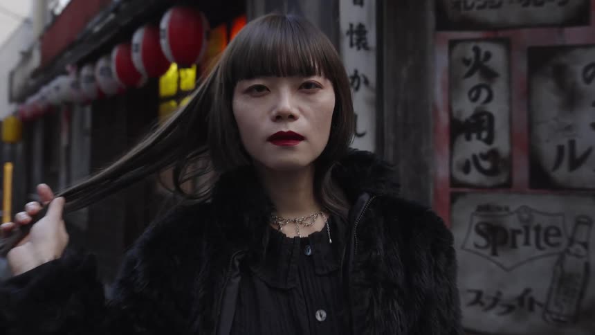 A stylish woman with bangs and red lipstick stares intensely at the camera while holding her hair in a Japanese alley, as the camera slowly zooms in