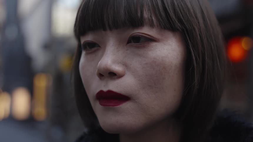 A close-up portrait of a pensive Asian woman with red lipstick and a bob haircut on a city street at night