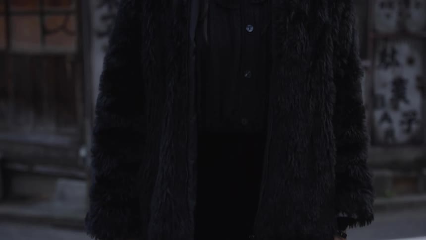 A slow tilt-up reveals a stylish Japanese woman in a black fur coat and red lipstick, staring intensely at the camera on an old city street